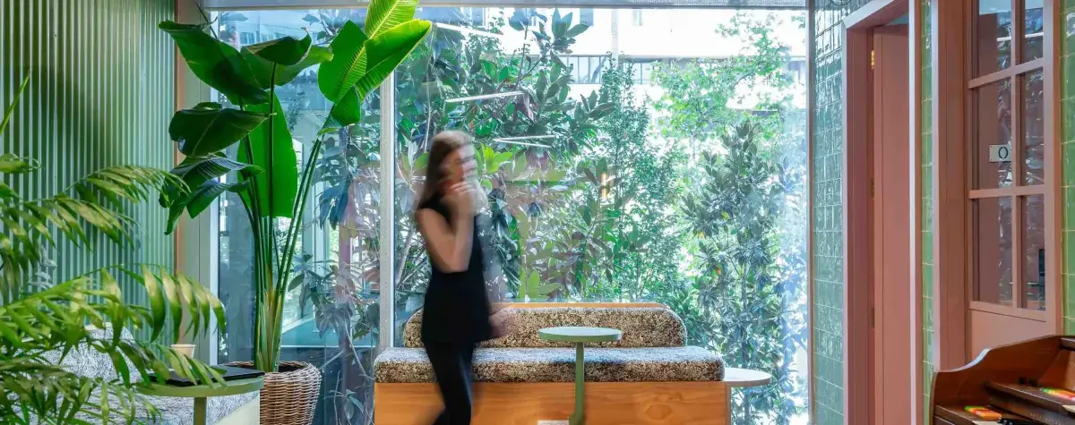 Una persona camina mientras habla por teléfono en un espacio interior moderno con plantas grandes, un sofá y paredes de vidrio que dejan ver un jardín exterior.