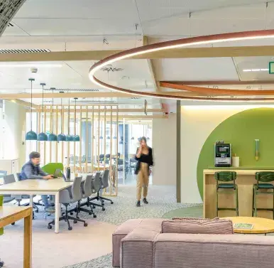 Un espacio de oficina moderno con personas trabajando y moviéndose. Hay mesas largas, sillas ergonómicas, una barra con taburetes y decoración circular en el techo. Ambiente luminoso y ordenado.