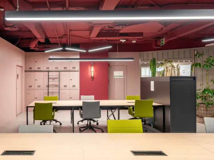 Oficina con sillas de colores alrededor de mesas largas; armarios y plantas en un entorno moderno con techo rojo.