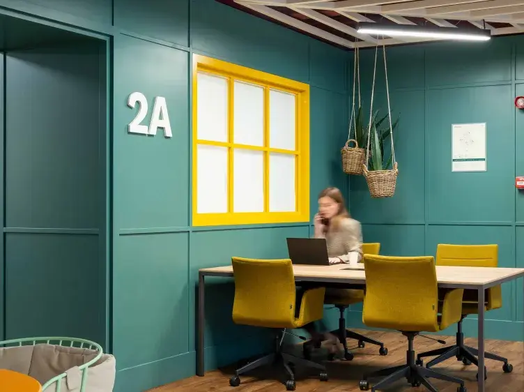 Una mujer trabaja en una mesa con computadora en una oficina moderna. En la pared, se lee "2A" junto a una ventana amarilla, con macetas colgantes al fondo.