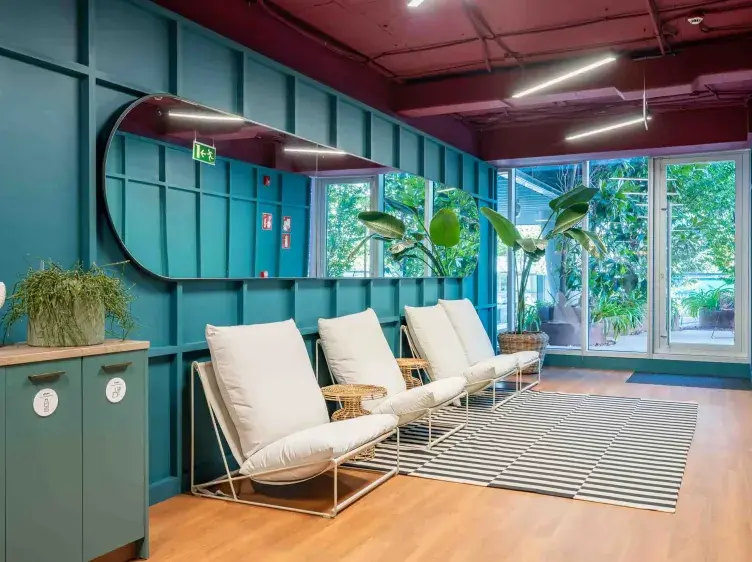 Sillas blancas descansan junto a una pared azul con espejo, en un espacio interior moderno con plantas y piso de madera. Hay un cartel de salida de emergencia iluminado.