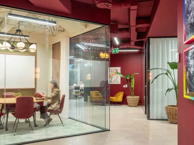 Una sala de reuniones con paredes de vidrio muestra a una persona sentada. Contexto de oficina moderna con paredes rojas y plantas decorativas.