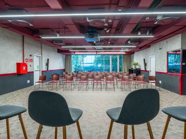 Un salón de conferencias con sillas alineadas, visto desde un podio. Al fondo, ventanas grandes ofrecen vista al exterior. Paredes blancas y detalles rojos complementan el ambiente.