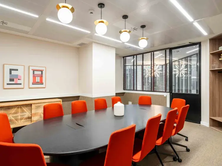 Una mesa ovalada negra rodeada de sillas naranjas en una sala de reuniones con lámparas colgantes esféricas. Hay cuadros en la pared y una puerta de vidrio con texto indescifrable al fondo.