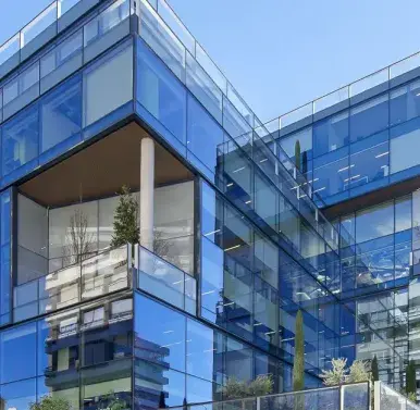 Un edificio moderno con fachada de vidrio refleja el cielo y sus alrededores; plantas decoran varias terrazas.