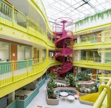 Una escalera roja en espiral conecta diferentes niveles en un edificio de oficinas con corredores amarillos, bajo un techo de vidrio y rodeado de plantas y áreas de descanso.