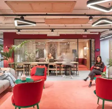 Un espacio moderno con sillones rojos y grises, personas conversan y trabajan en un entorno luminoso. Decoración contemporánea y plantas. Letreros visibles: "SALIDA" y un panel con pantalla iluminada.