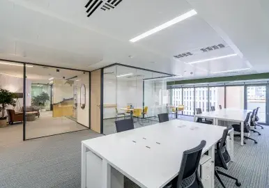 Una oficina moderna con escritorios blancos y sillas negras está vacía. Ventanas amplias permiten la entrada de luz natural. Una sala de reuniones se ve a través de un muro de vidrio.