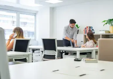 Un hombre señala una computadora portátil sobre una mesa mientras una mujer lo observa, en una oficina con libros y plantas.